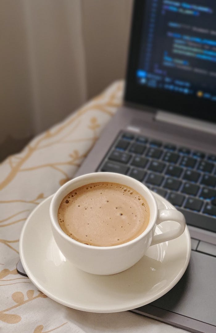 pexels-photo-31340326 Cup of frothy coffee on a laptop keyboard with coding screen, cozy indoor setting.